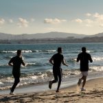 Three men jogging along a scenic beach, embracing a healthy lifestyle under a clear sky.